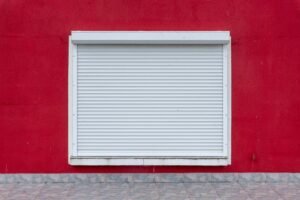 Volet roulant blanc entièrement descendu sur une fenêtre, coffre en applique visible au-dessus, mur rouge