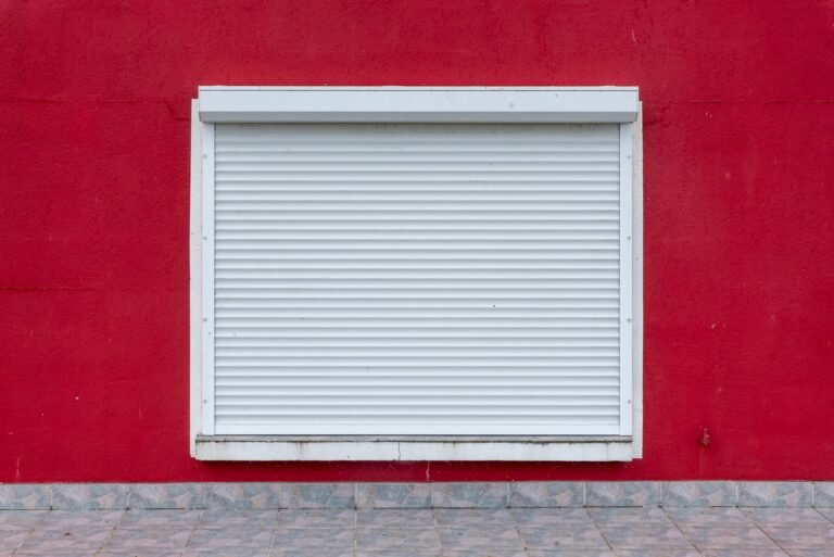 Volet roulant blanc entièrement descendu sur une fenêtre, coffre en applique visible au-dessus, mur rouge
