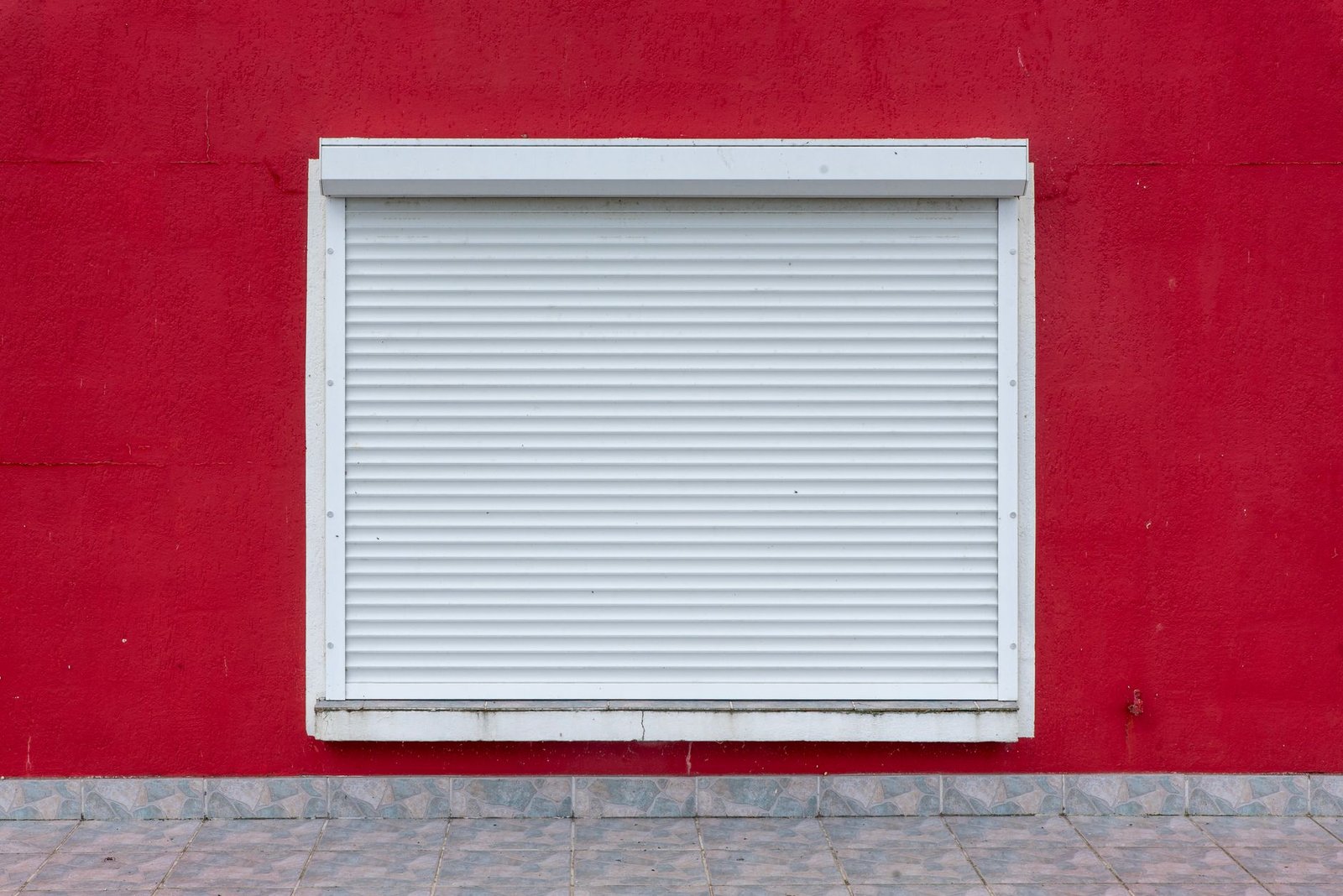 Volet roulant blanc entièrement descendu sur une fenêtre, coffre en applique visible au-dessus, mur rouge