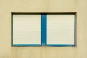 Fenêtre à deux vantaux fermée par des volets roulants crème, encadrement bleu sur une façade beige