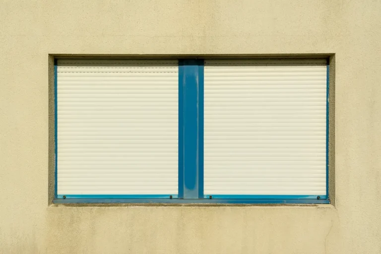 Fenêtre à deux vantaux fermée par des volets roulants crème, encadrement bleu sur une façade beige